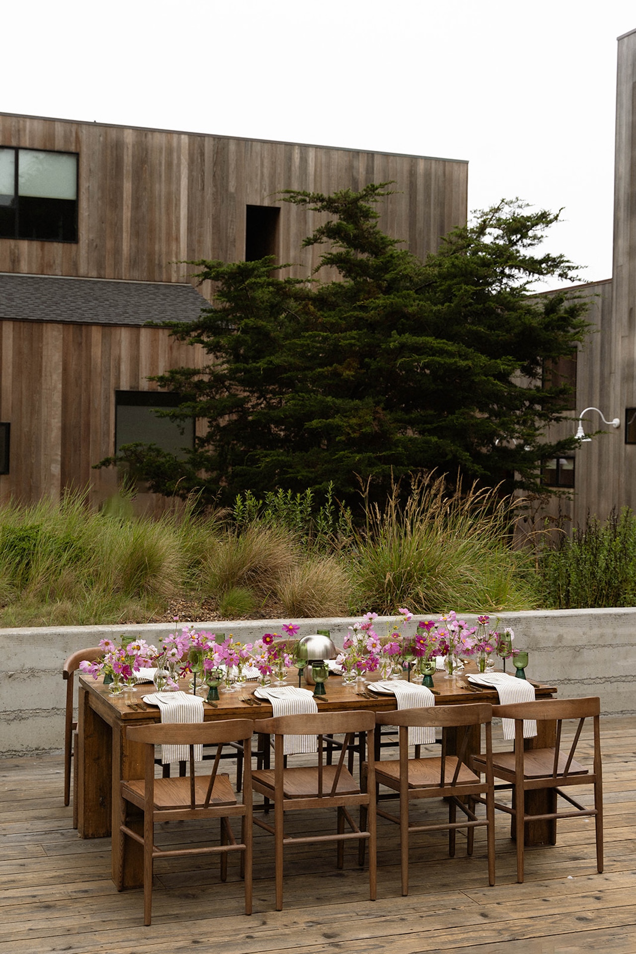 Wide view of a long wooden dining table surrounded by greenery and modern architecture, styled for an intimate outdoor celebration.