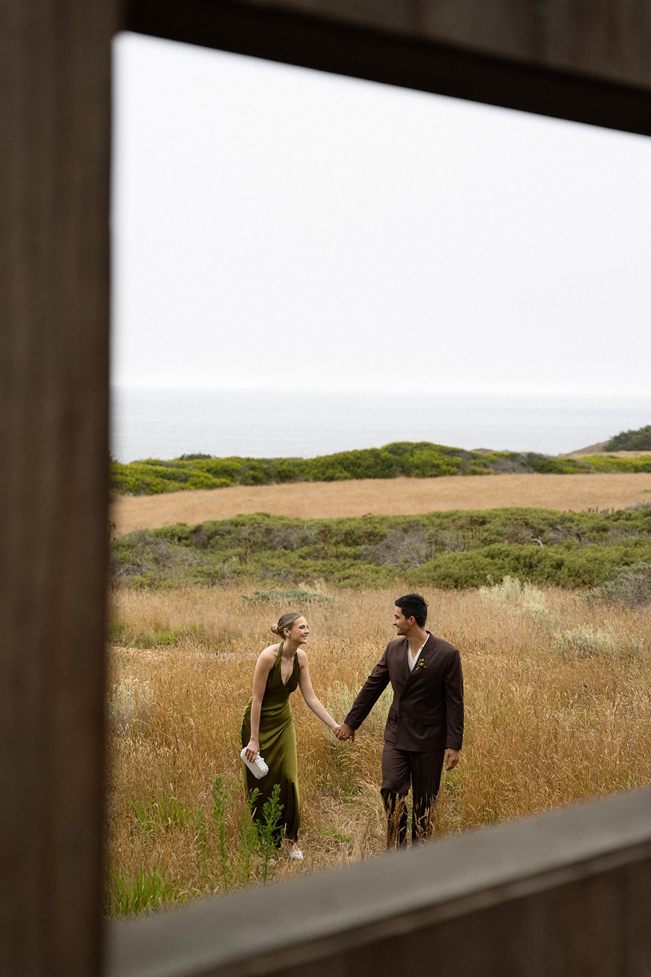 Film-style portrait of the couple standing together in tall grass, framed by rolling hills and muted coastal tones.