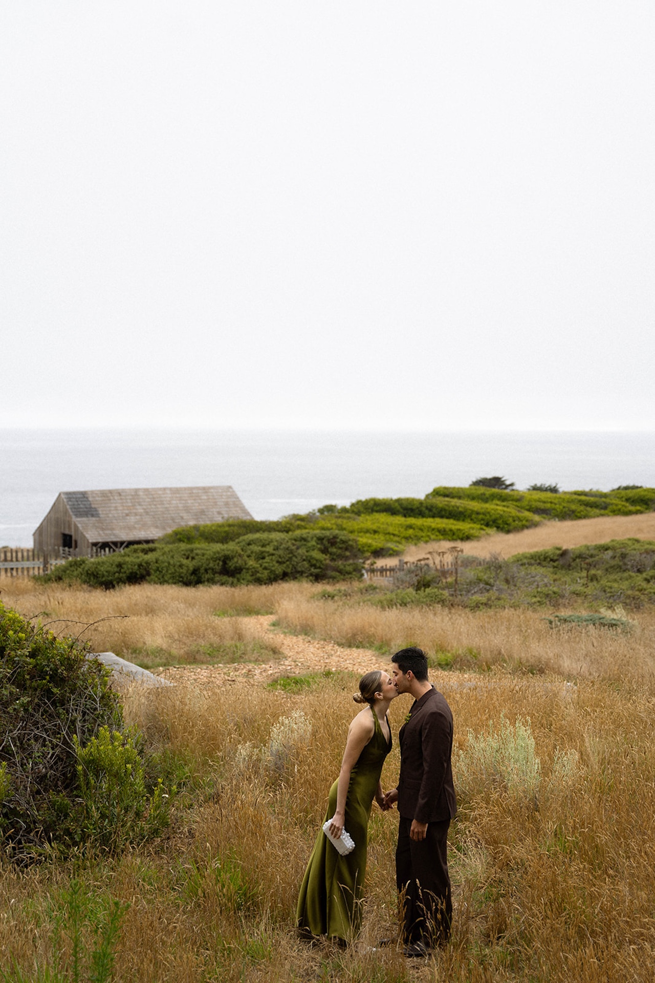 Bride and groom kissing in tall grass with rolling coastal hills in the background, reflecting intimacy and intention while planning a destination wedding.
