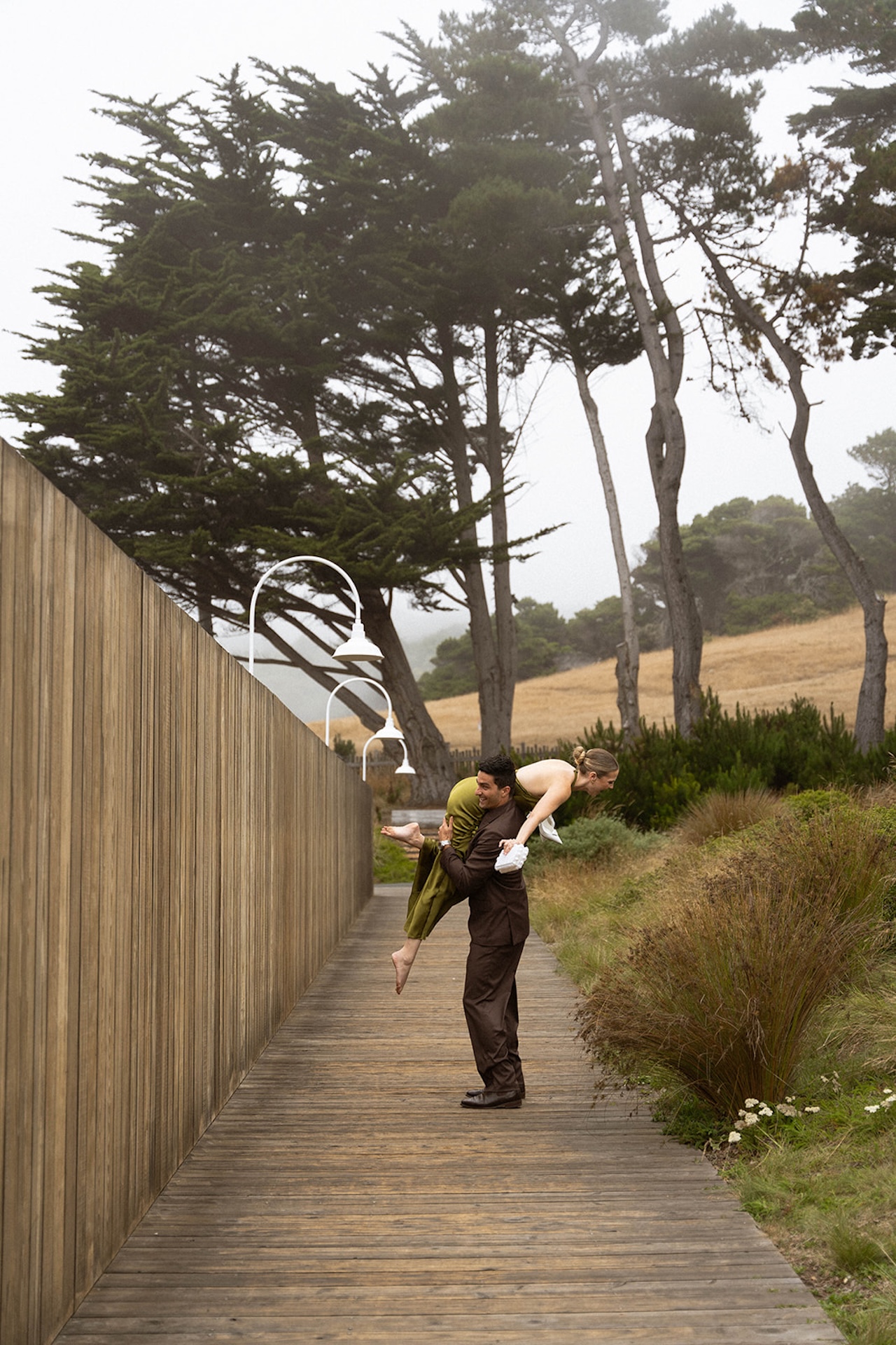 Couple laughing on a wooden boardwalk as the groom lifts the bride, surrounded by coastal grasses and tall trees.