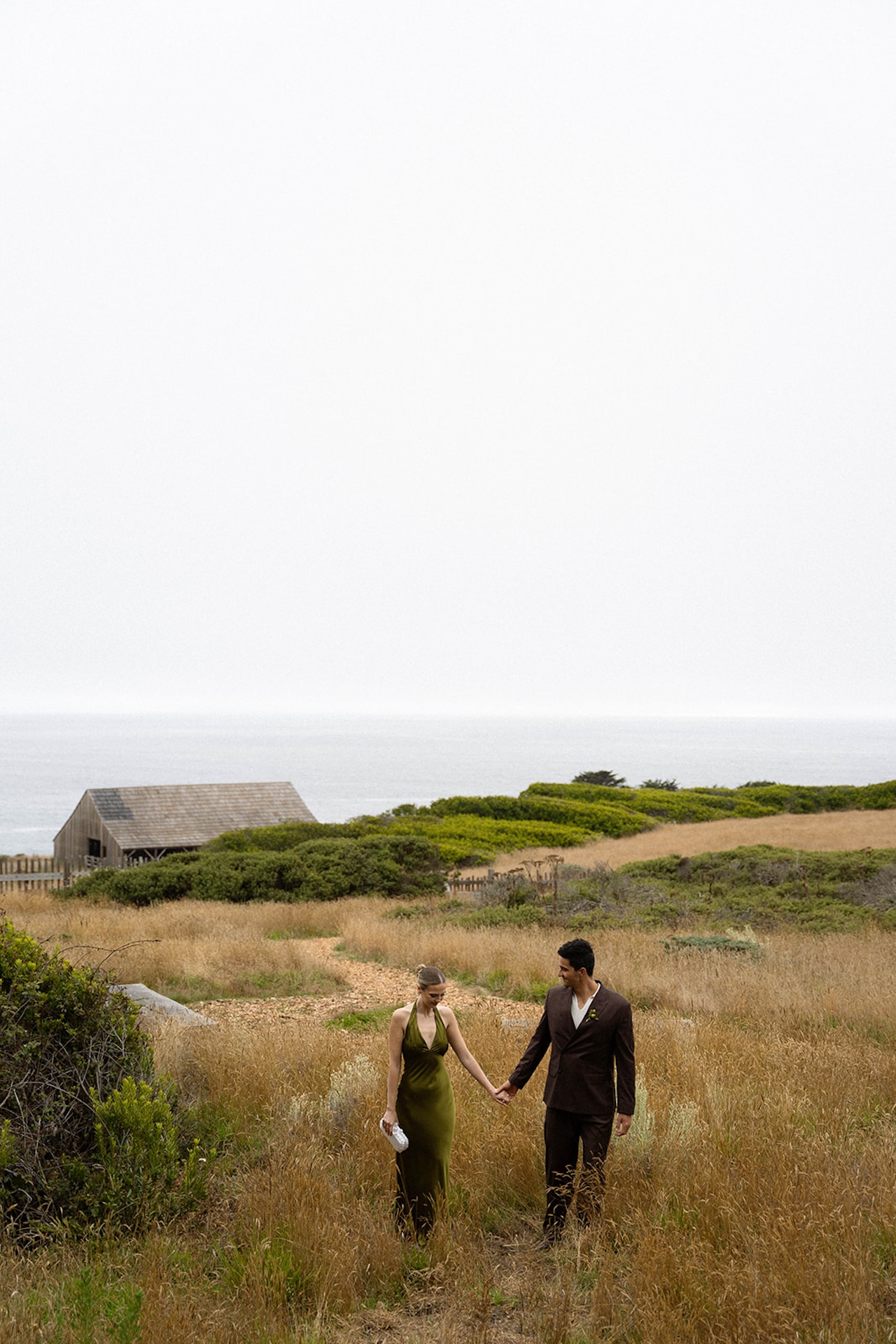 Bride and groom walking hand in hand through tall coastal grass with the ocean behind them, embracing a slow moment while planning a destination wedding.