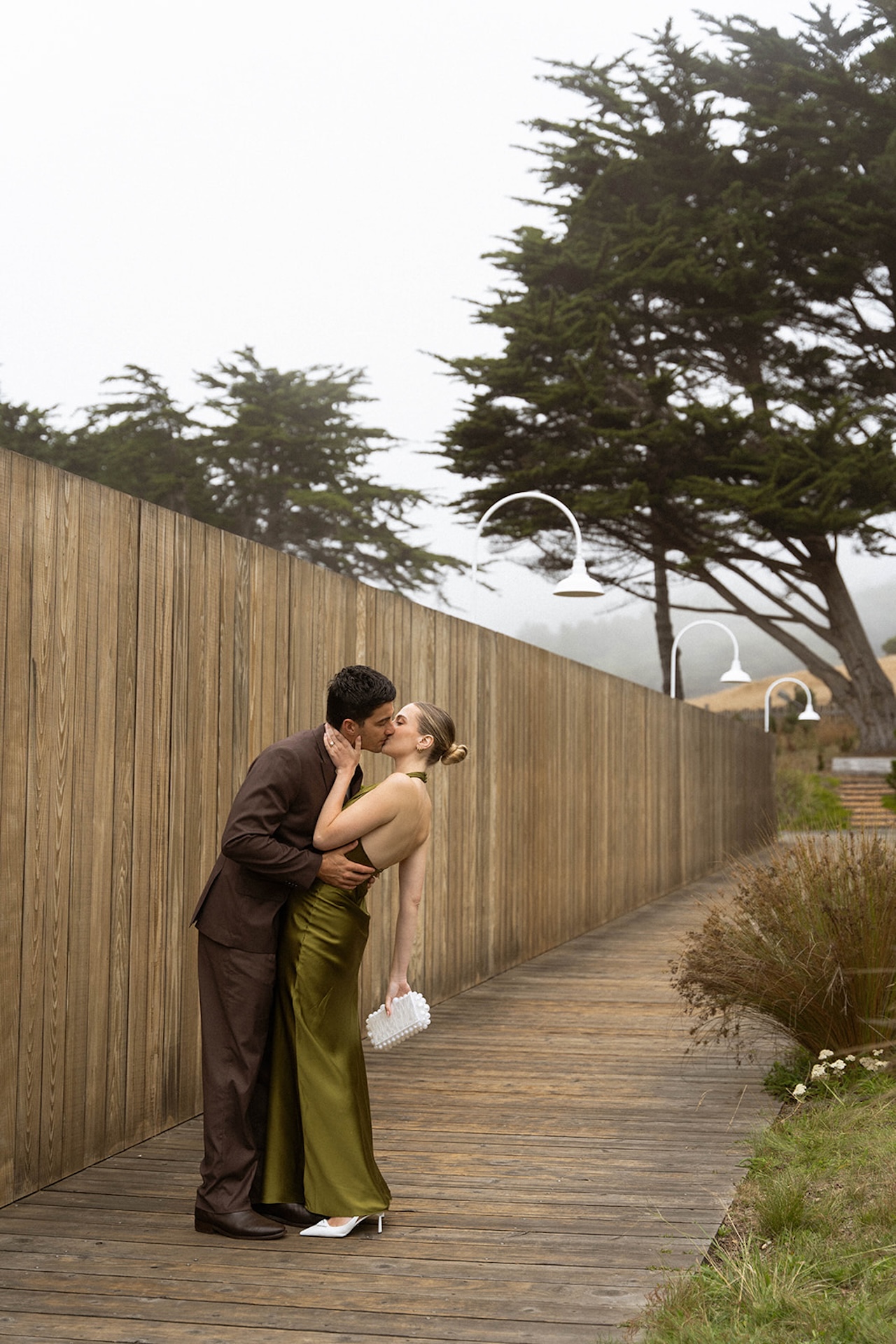 Bride and groom sharing a quiet kiss on a wood walkway surrounded by foggy coastal greenery, captured in a soft editorial style.