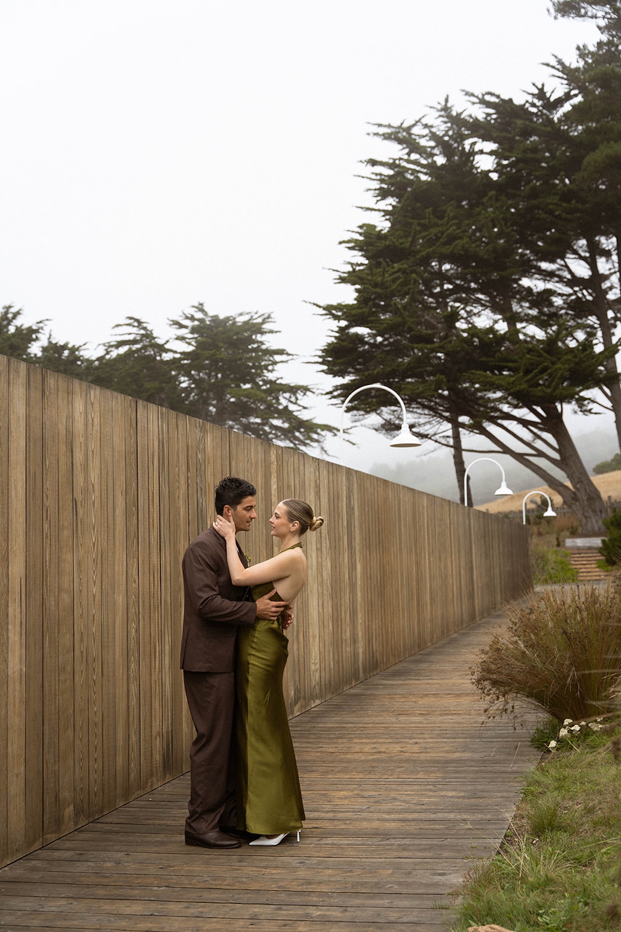 Bride and groom standing close on a wooden boardwalk with coastal trees in the background, wearing modern olive and brown wedding attire.