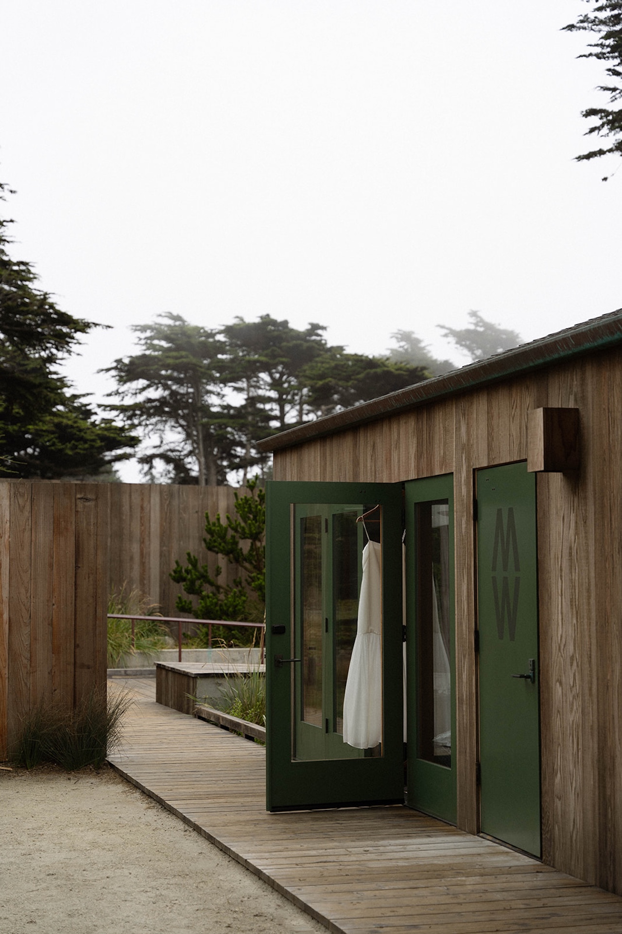 Modern wooden cabin exterior with green doors and a white wedding dress hanging inside, set along a coastal boardwalk.