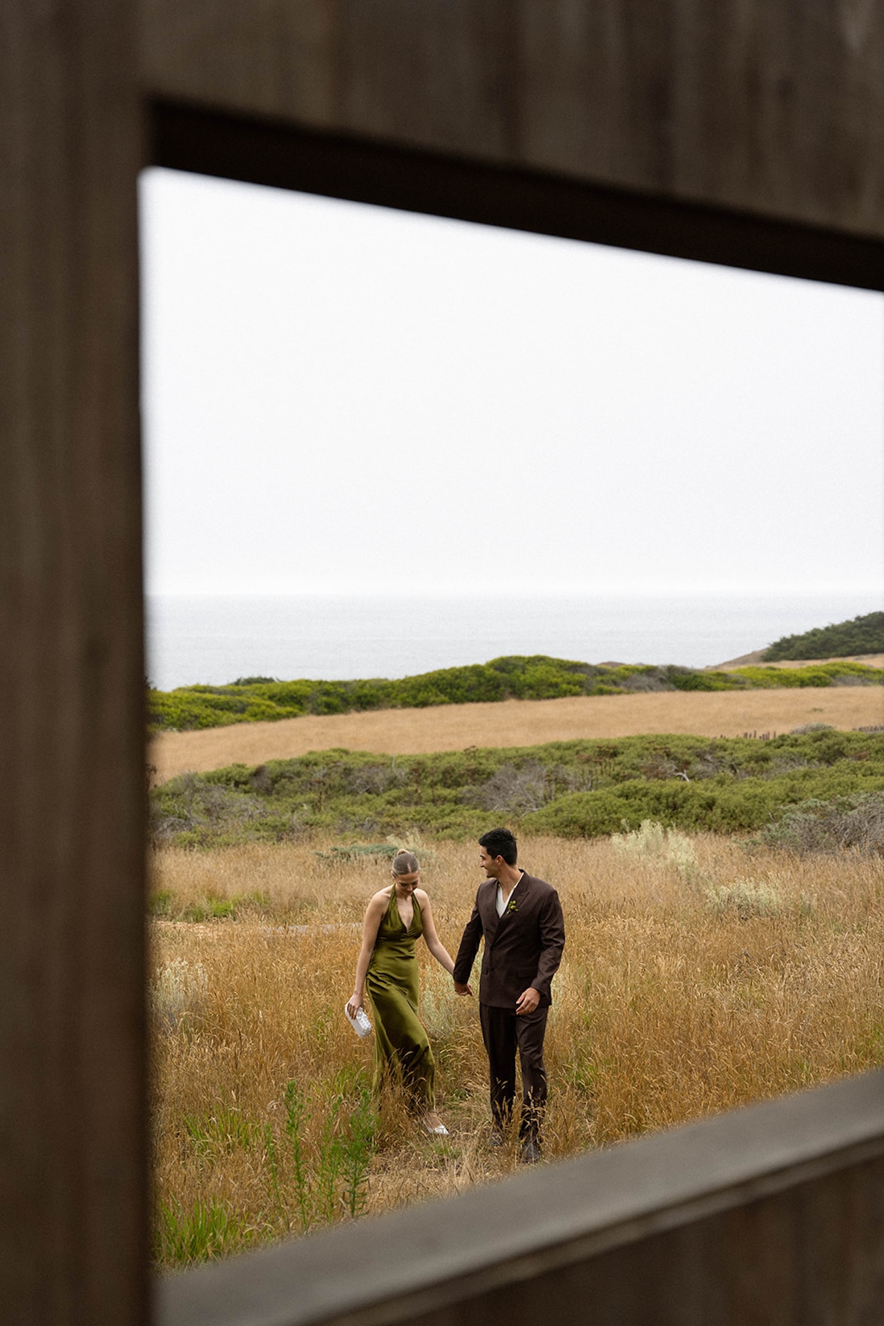 Wide editorial shot of the couple standing together in a golden coastal field with rustic buildings and soft ocean light.