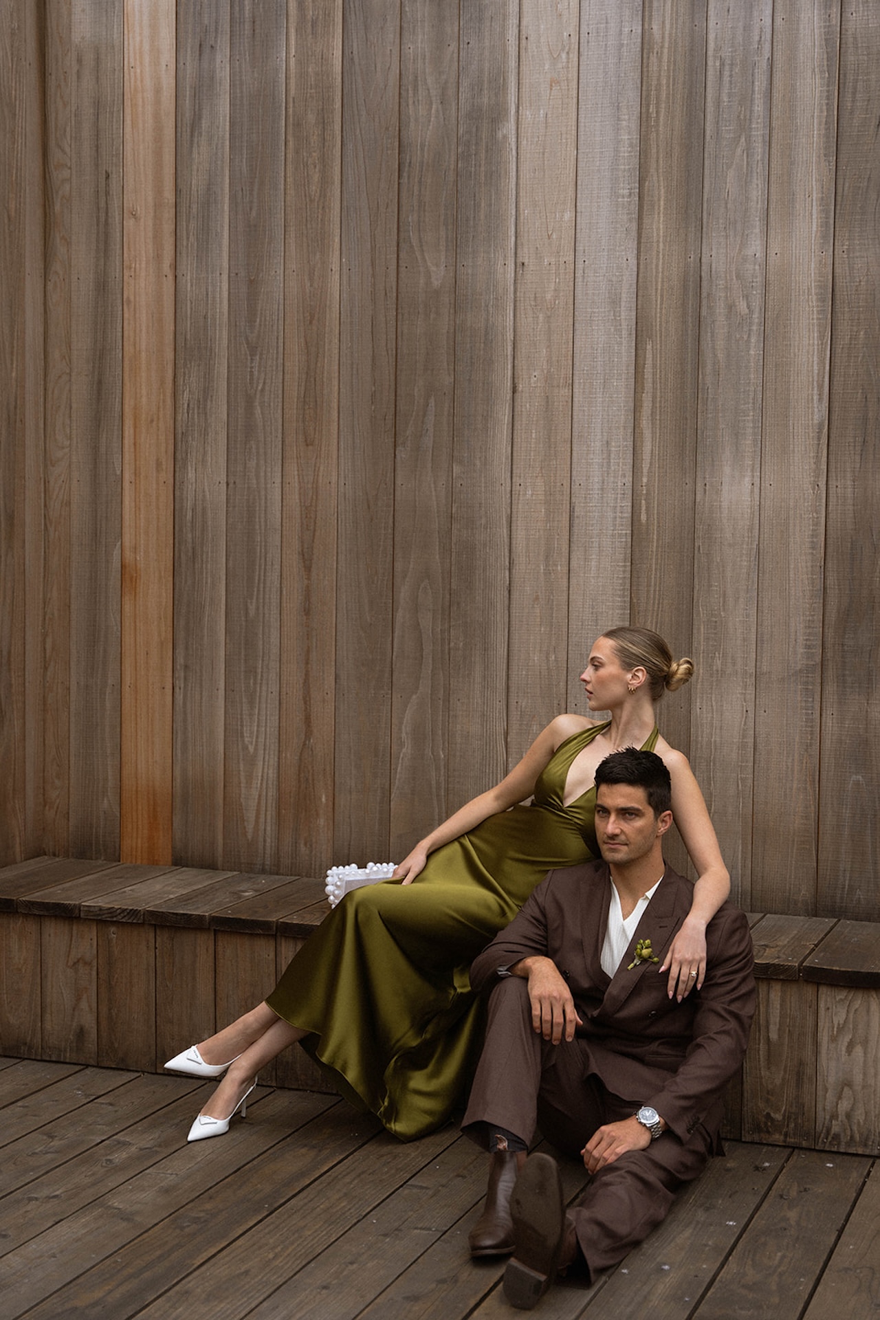 Bride in an olive green halter dress and groom in a brown suit sitting on a wooden deck against a modern wood wall, relaxed and editorial in style.