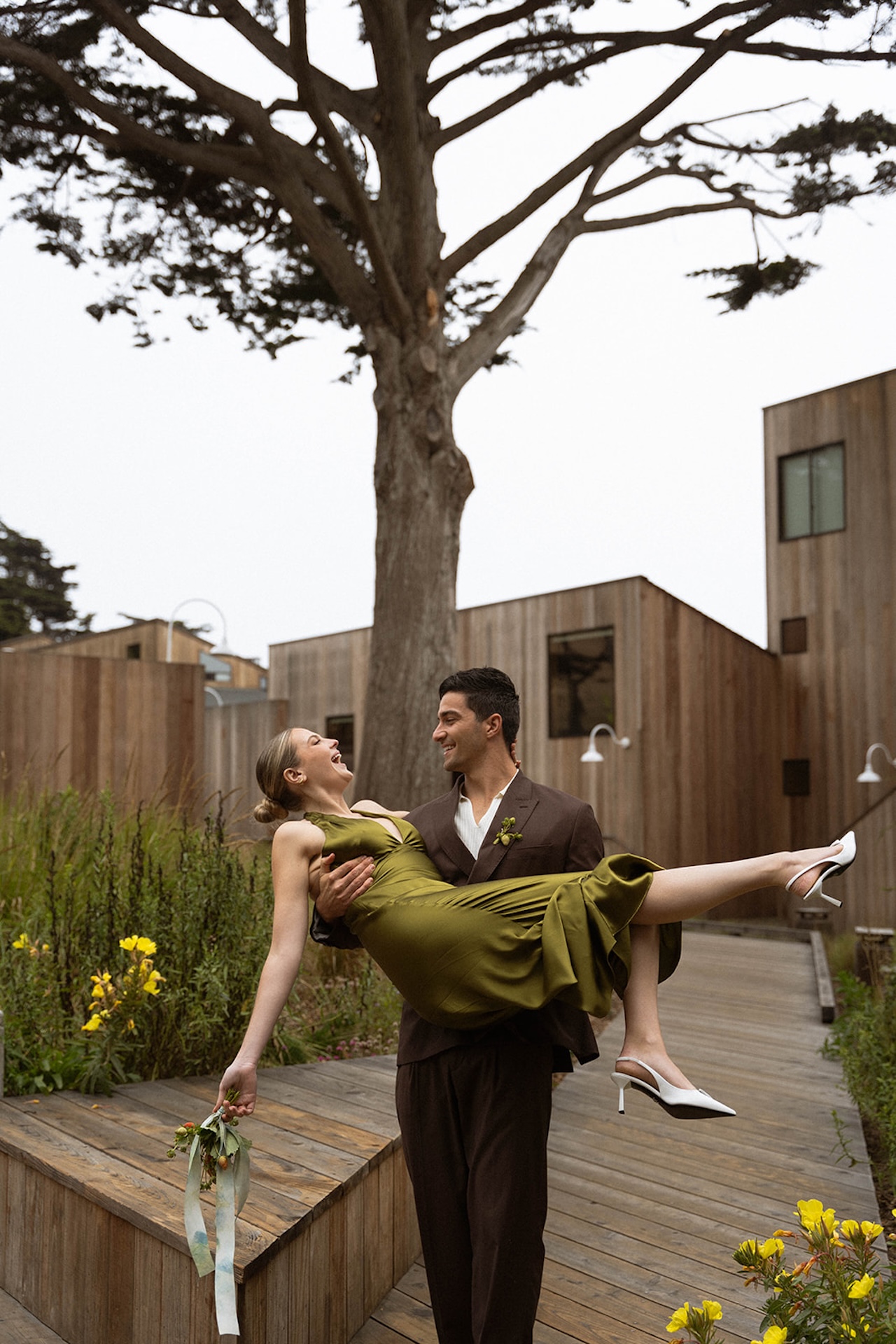 Bride and groom laughing as the groom carries the bride along a wooden path between modern cabins, ending the day with playful energy.