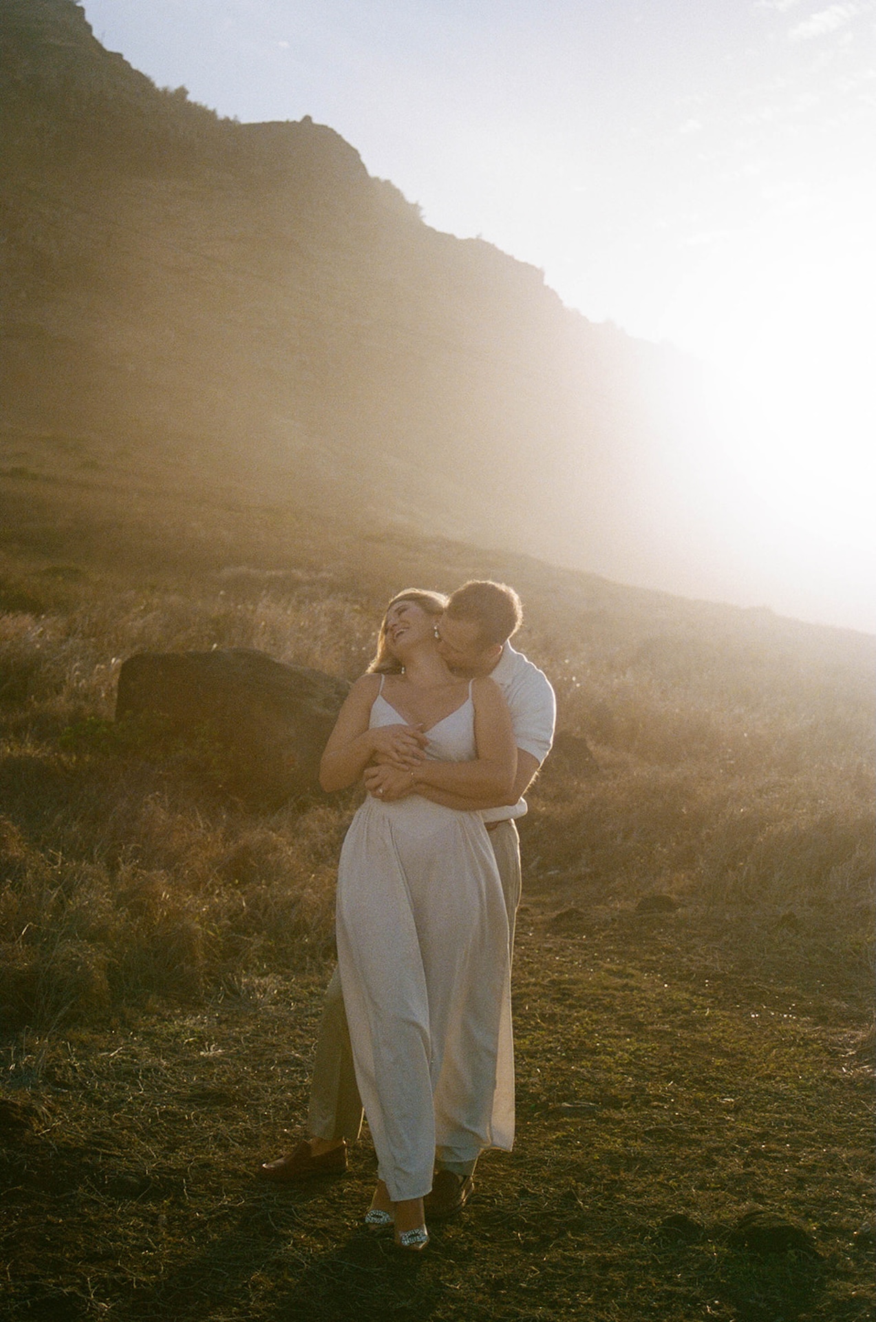 A couple standing in tall grass at golden hour, wrapped in each other’s arms with soft sunlight and wind-blown movement.