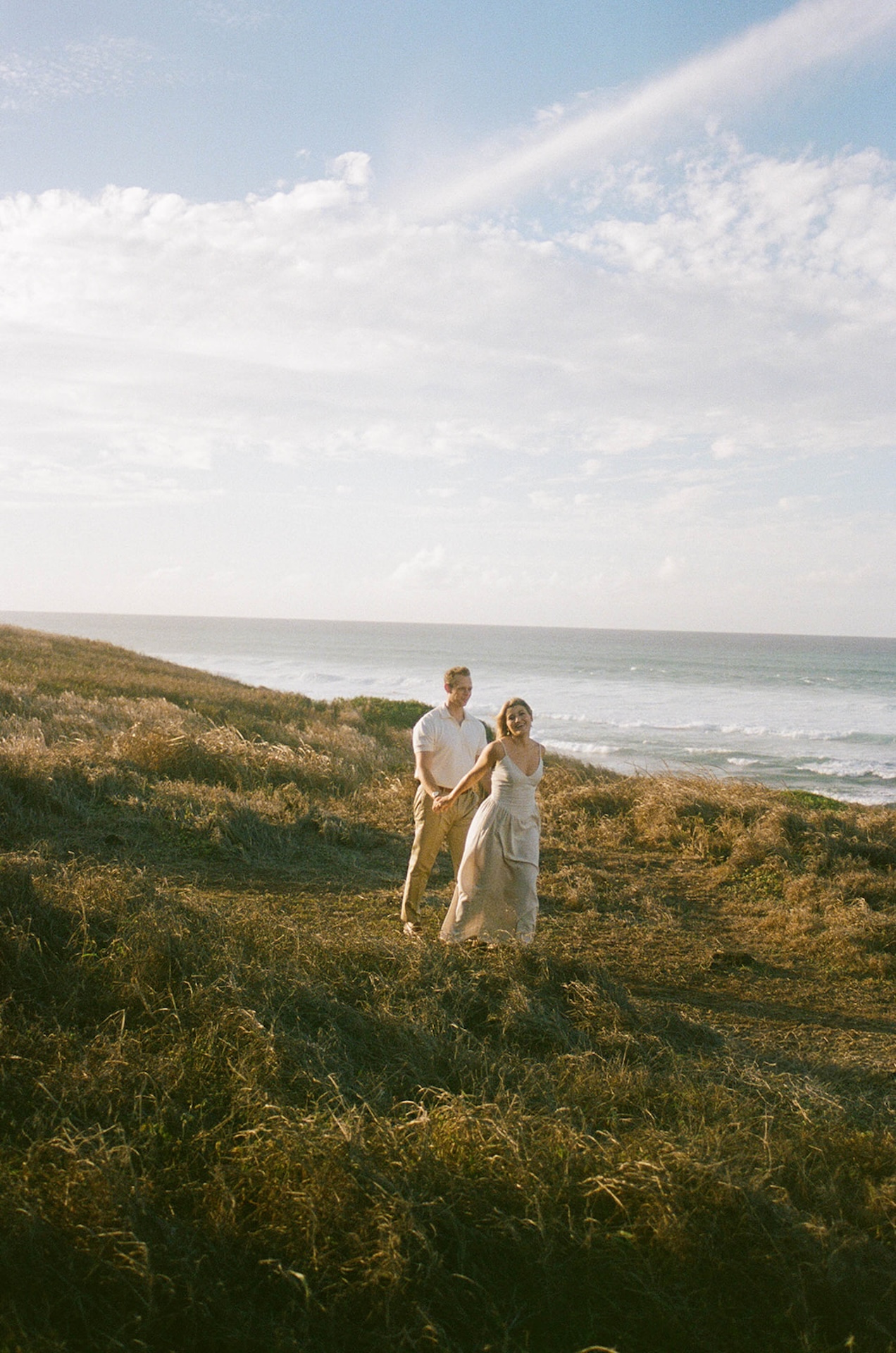 The couple walking hand in hand through windswept grass with the ocean stretching behind them under a bright, open sky.