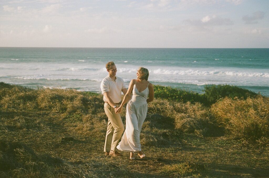 A playful walking moment near the coastline as the couple laughs together with waves crashing in the background.