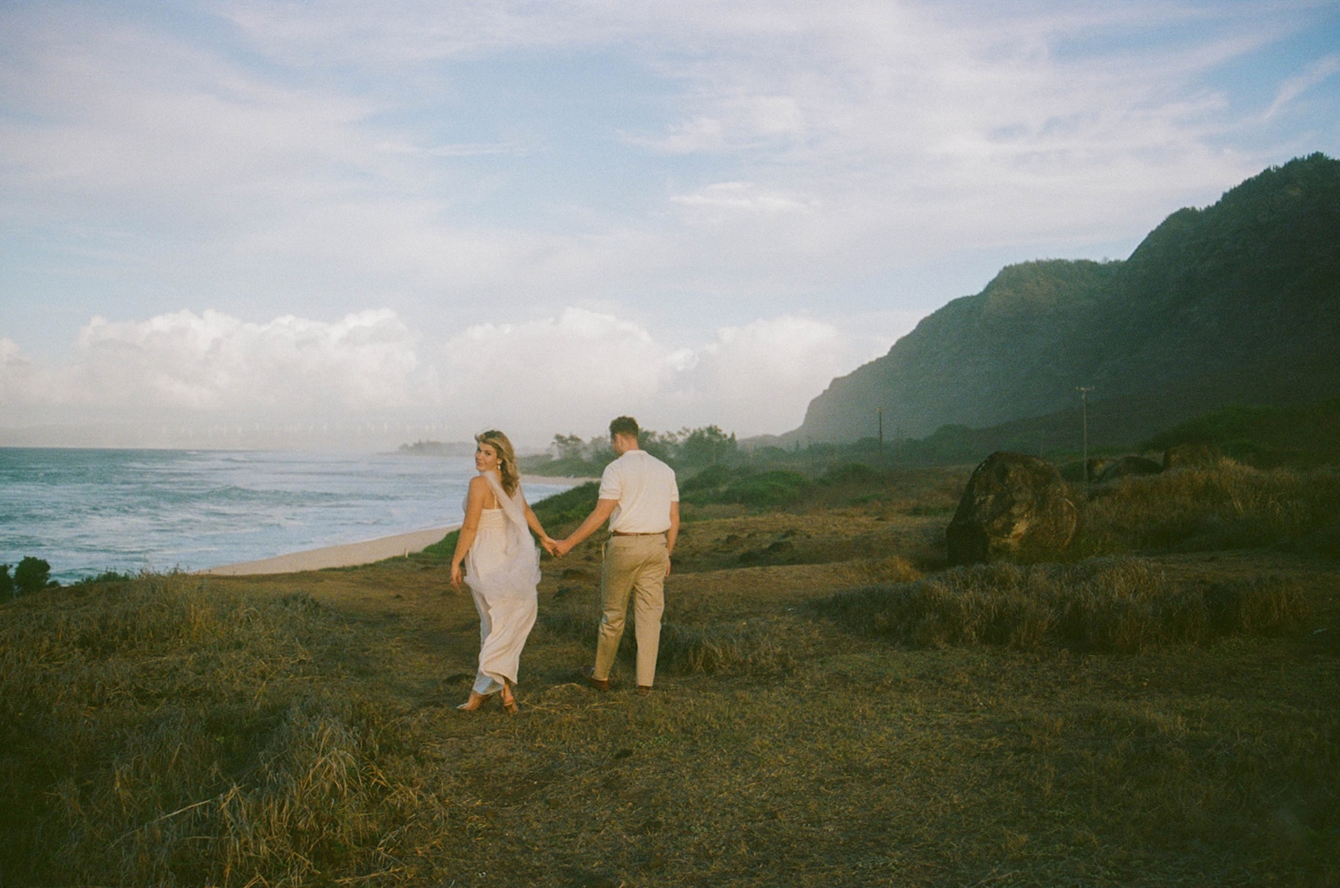The couple holding hands and walking along a grassy coastal path with cliffs and ocean views in the background.