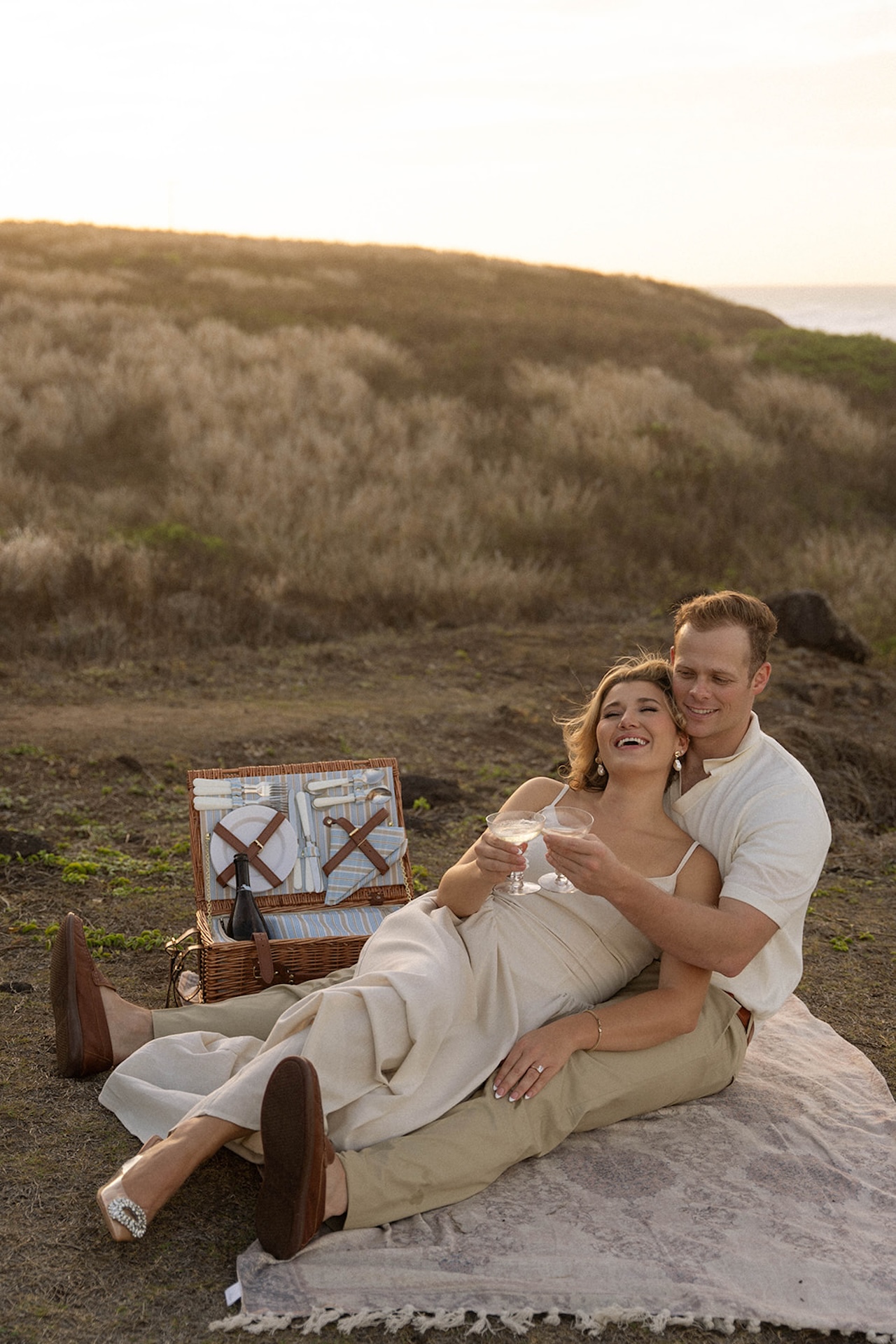 A relaxed picnic-style engagement moment with a couple sitting on a blanket, clinking glasses near the ocean, showcasing casual coastal ideas for engagement photos