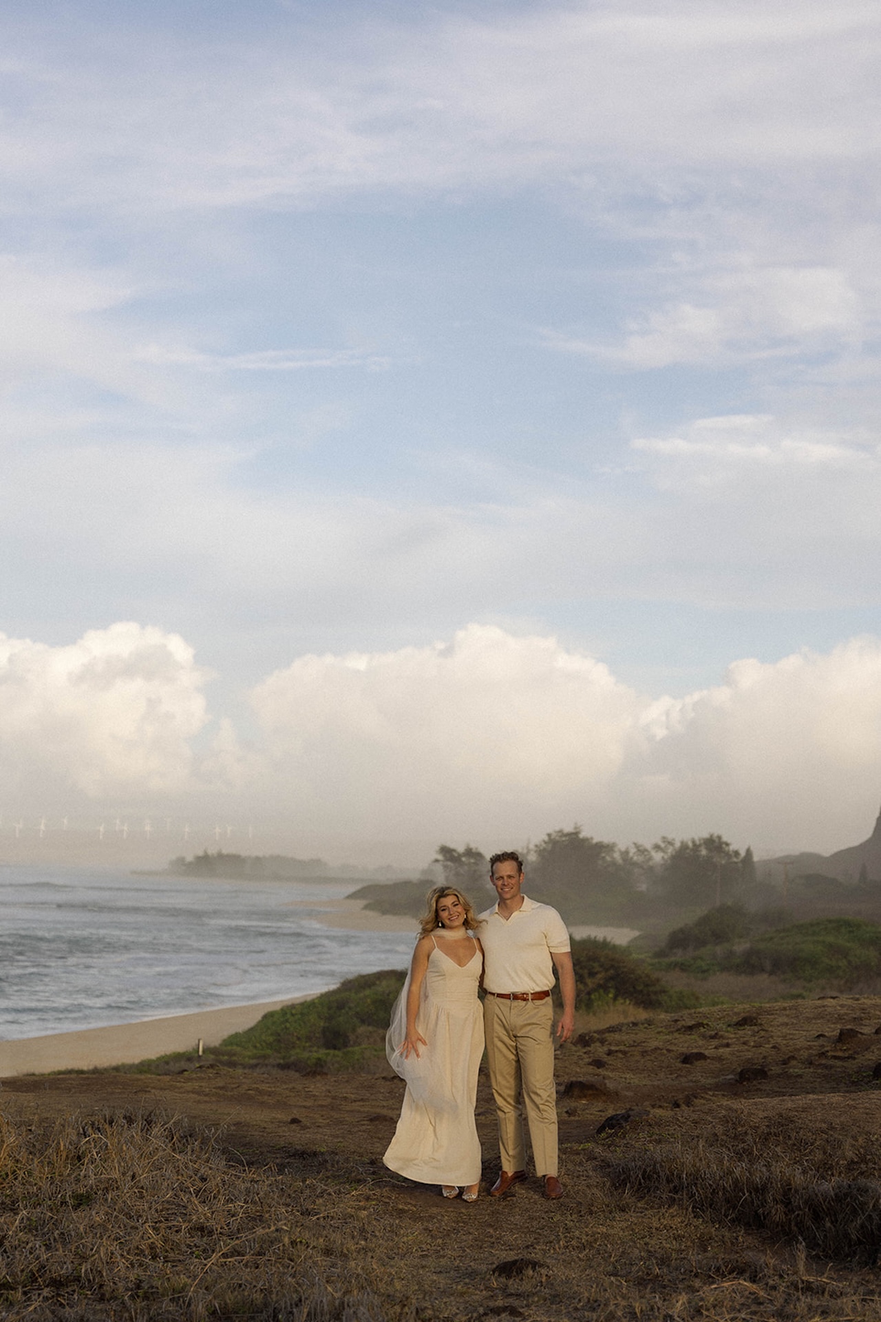 The couple standing close together with the ocean and cliffs in the background, sharing a quiet, candid moment.