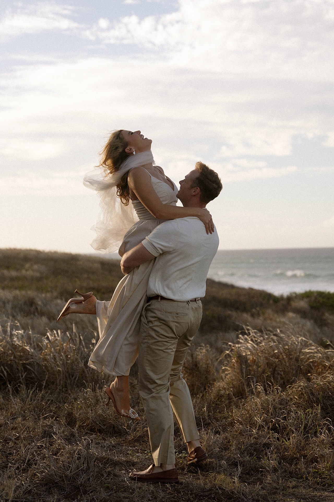 A playful lift as the couple laughs together in an open coastal field, showing joyful ideas for engagement photos on oahu.