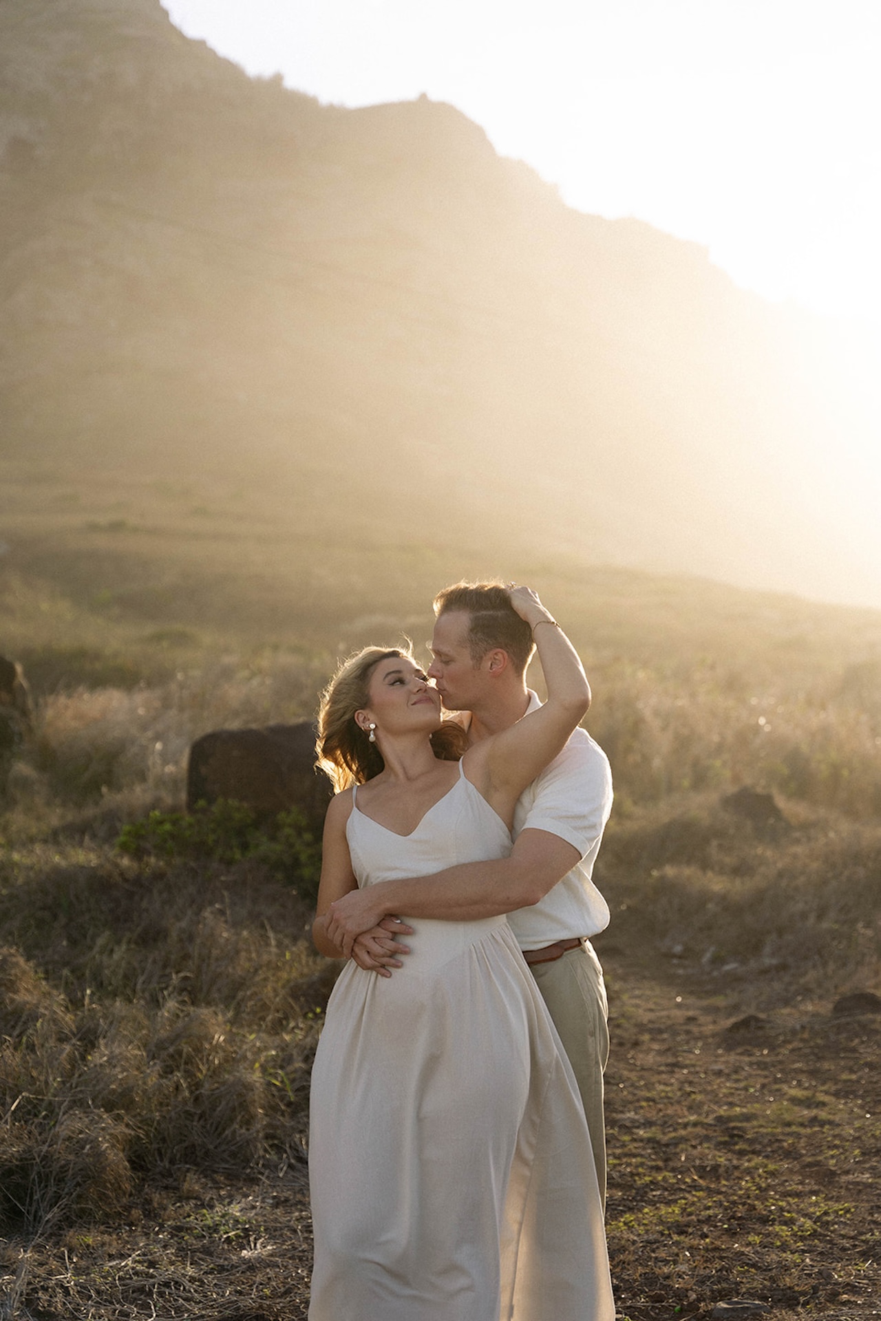 A backlit embrace in an open field as the couple stands close together, framed by soft golden light and rugged hillside textures.