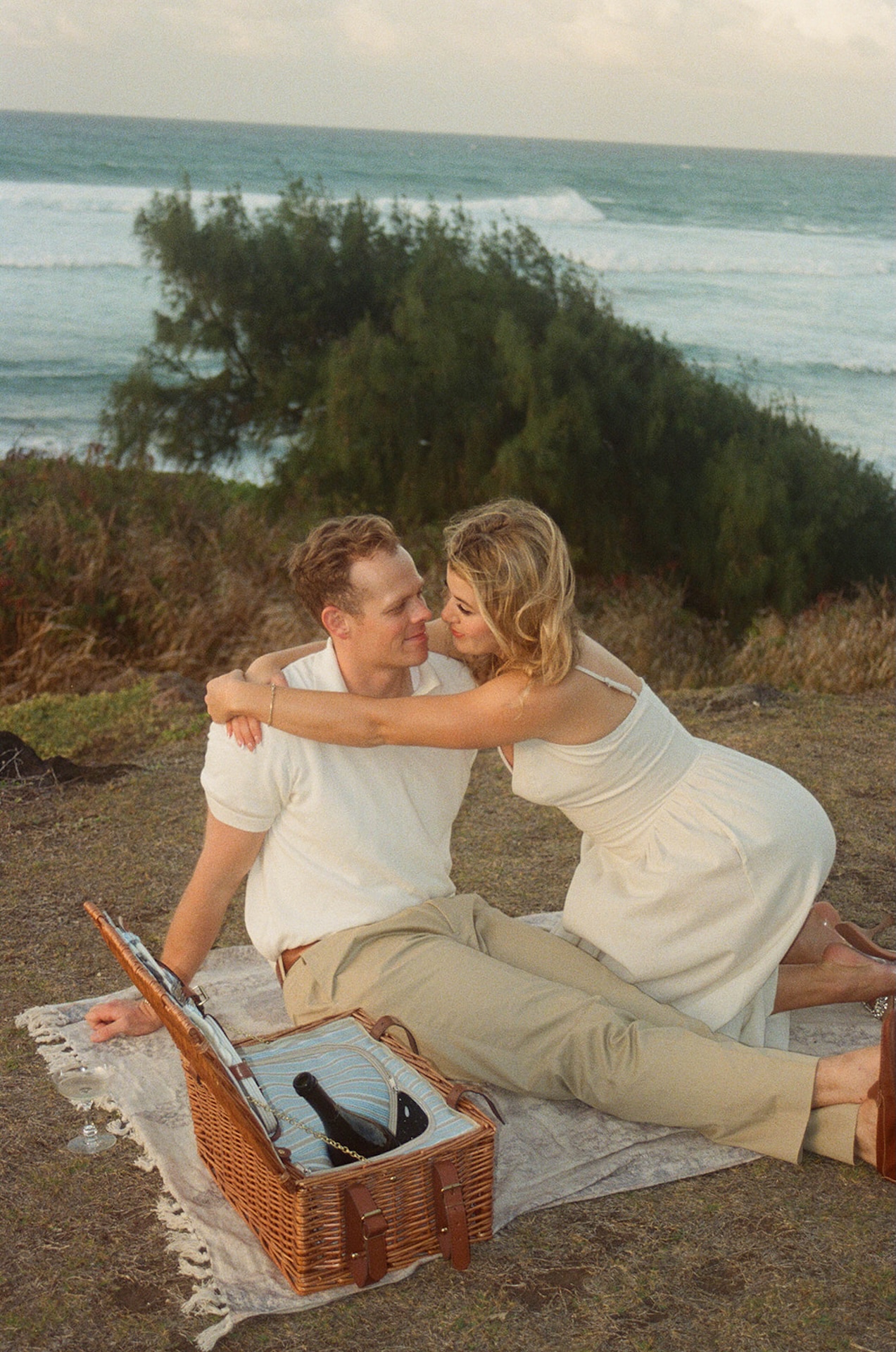 A cozy picnic moment as the couple sits facing each other on a blanket, sharing a quiet laugh with champagne nearby.