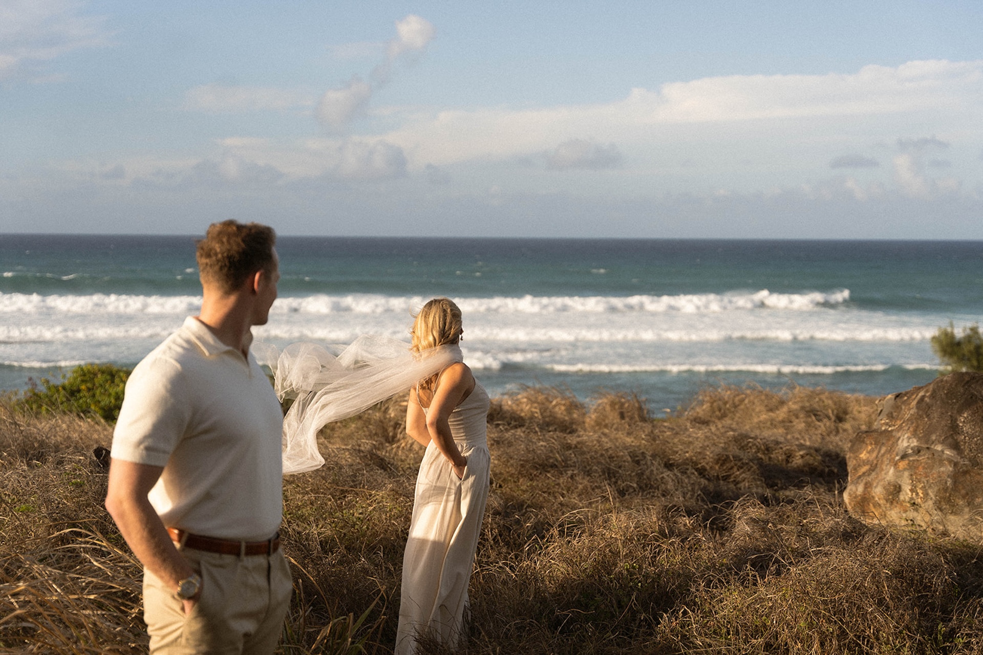The couple pausing along a grassy cliffside as wind moves through the landscape and waves roll in below.