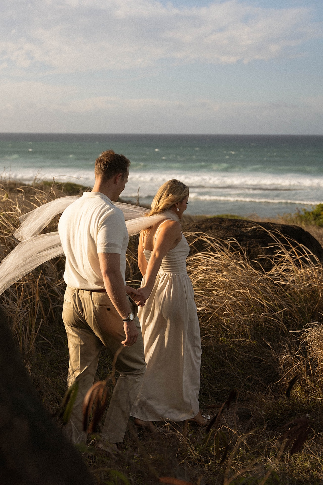 The couple pausing along a grassy cliffside as wind moves through the landscape and waves roll in below.