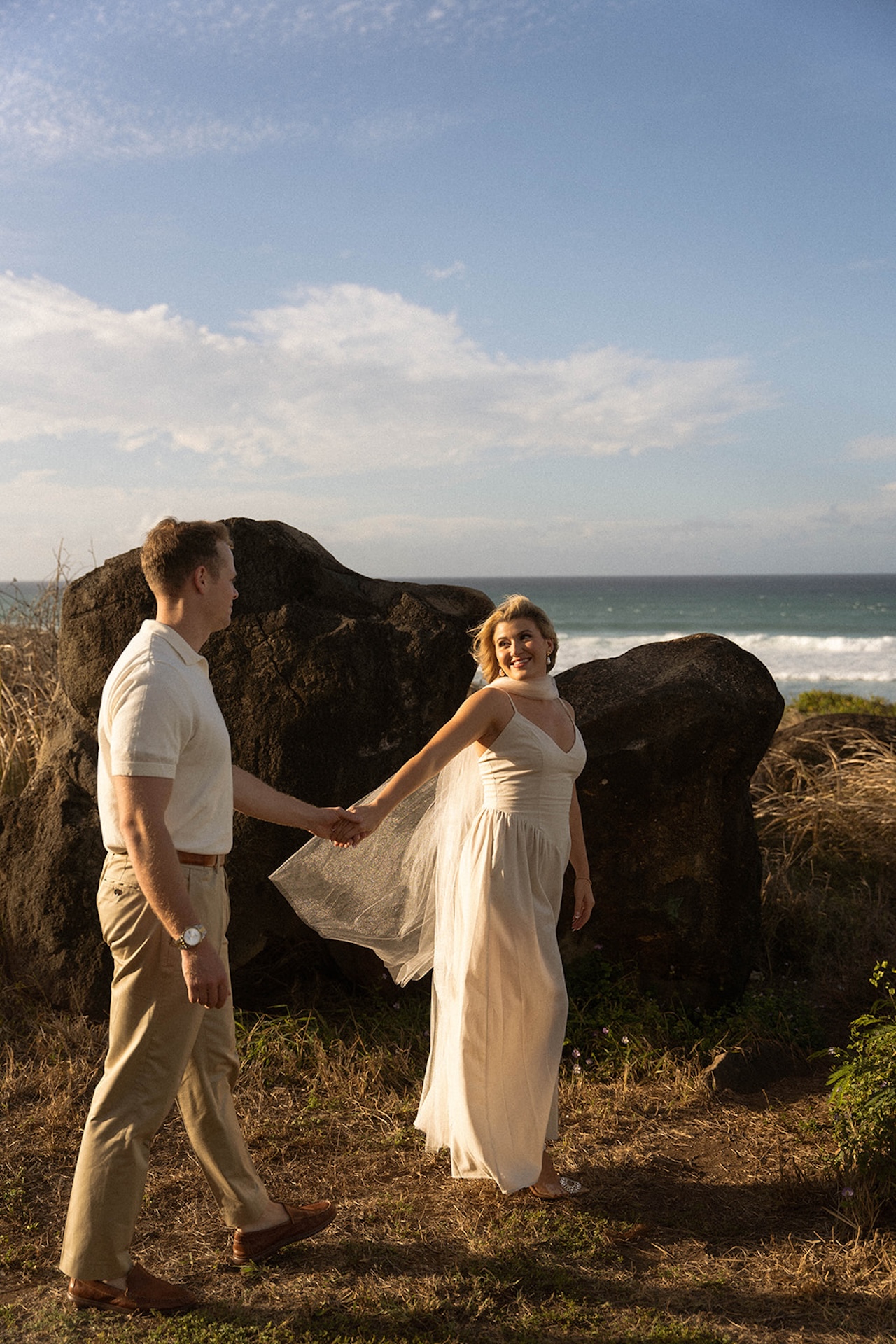 A couple holding hands while walking through a coastal field with the ocean behind them, highlighting relaxed and romantic ideas for engagement photos on oahu.
