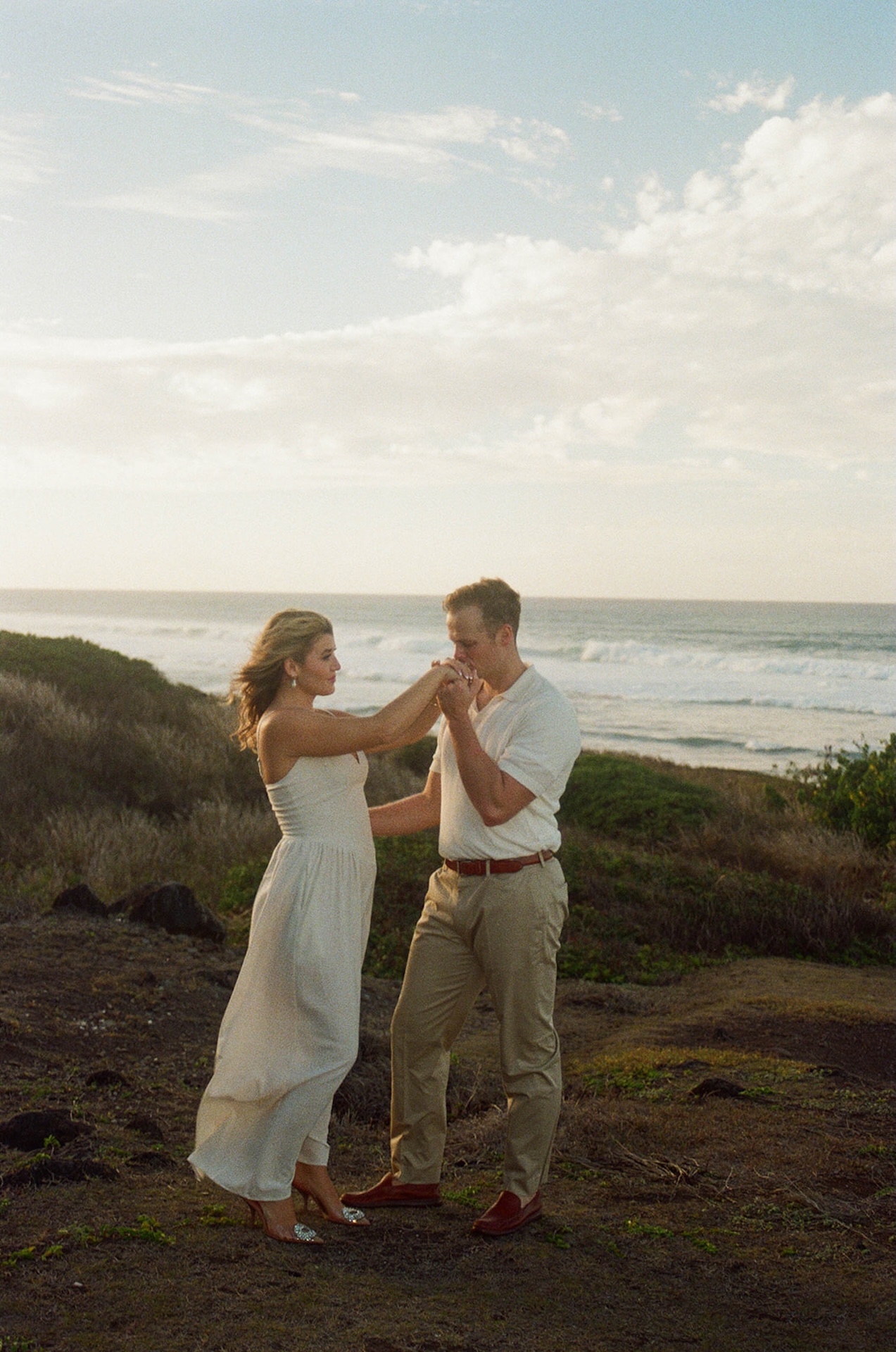 A couple standing together on a coastal bluff as one partner kisses the other’s hand, with ocean waves behind them, highlighting intimate ideas for engagement photos on oahu.
