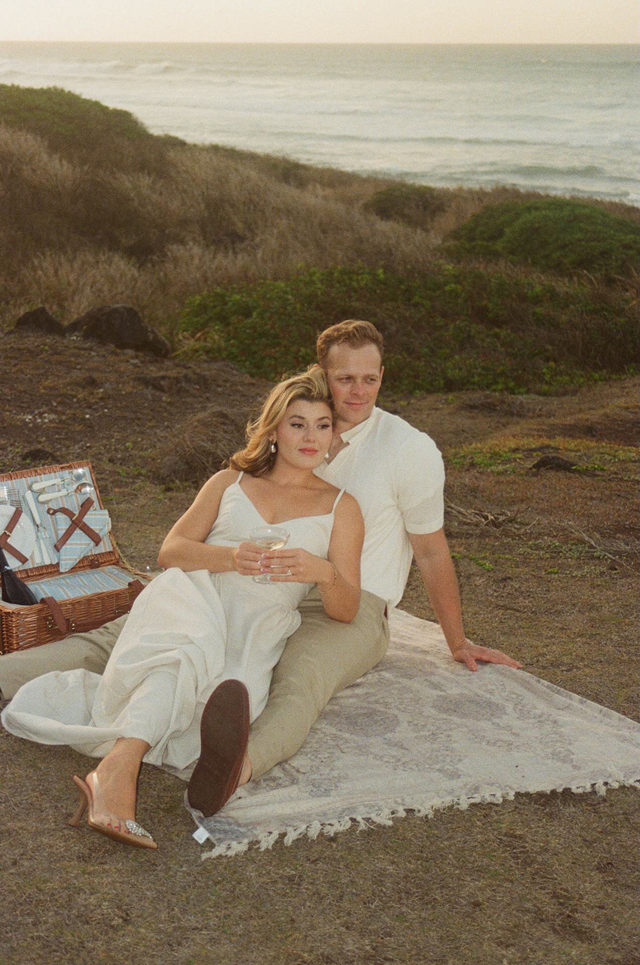 The couple sitting together on a picnic blanket near the ocean, relaxed and leaning into each other, showcasing lifestyle-inspired ideas for engagement photos on oahu.