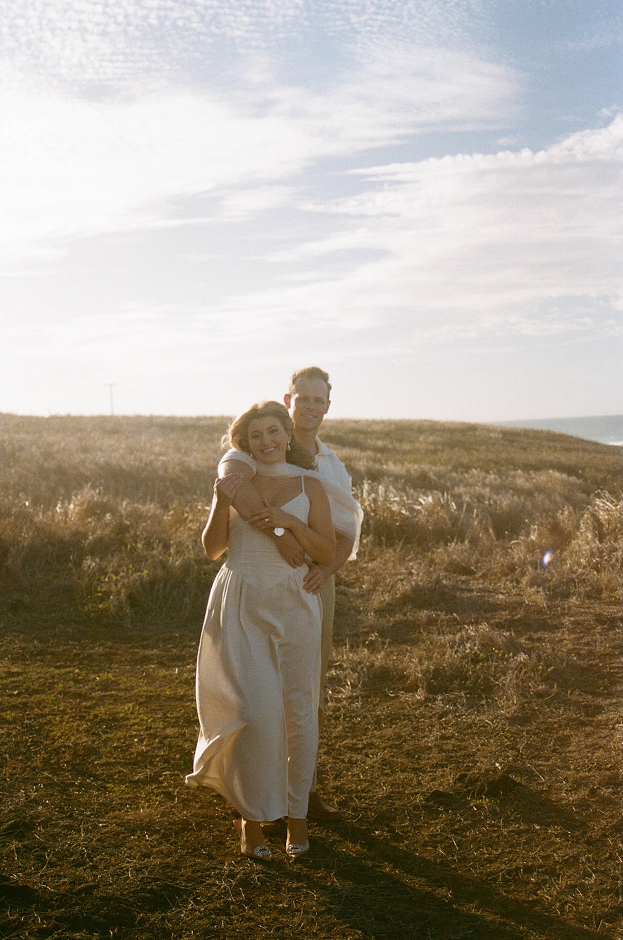 A cozy embrace with the groom standing behind the bride in a sunlit field, highlighting classic and intimate ideas for engagement photos on oahu.