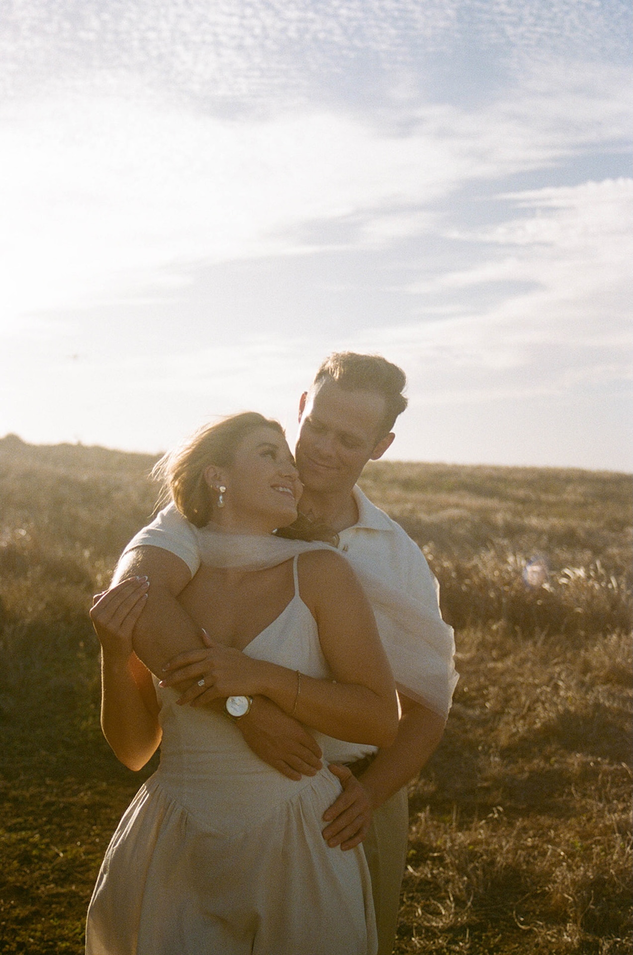 A softly backlit portrait of the couple standing together with rolling hills and glowing sunset light behind them.